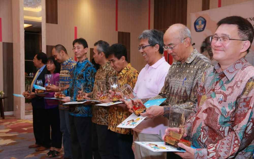 A group of people stands in a row at an indoor event, each holding a clear trophy and a colorful certificate or booklet. They are dressed in traditional and semi-formal batik shirts, with warm expressions on their faces, suggesting a recognition or award.
